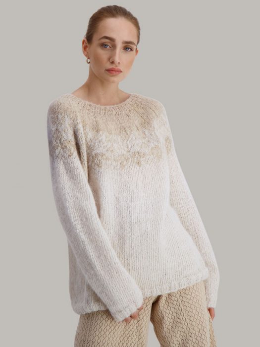 Strikkepakken Flora genser fra Skappel Classic. Bildet viser modellen i halvfigur. Modellen står i front og ser rett inn i kamera. Begge hendene henger ned langs sidene. Fargen på genseren er naturhvit og sandmelert.