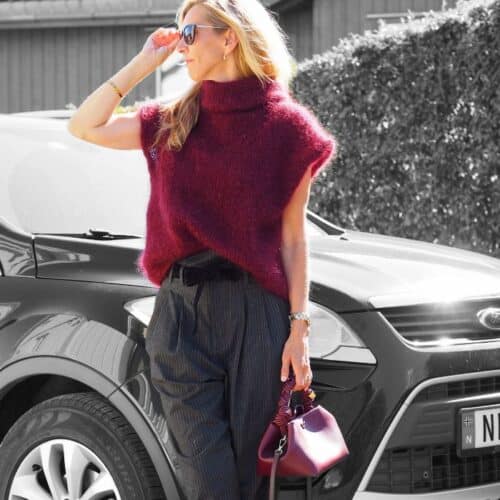 Produktbilde: Modell lett vridd til høyre. Står foran en bil. Strikkepakke Bea vest designet av Tove Lindtein, strikket i Bellissima fra by Permin.