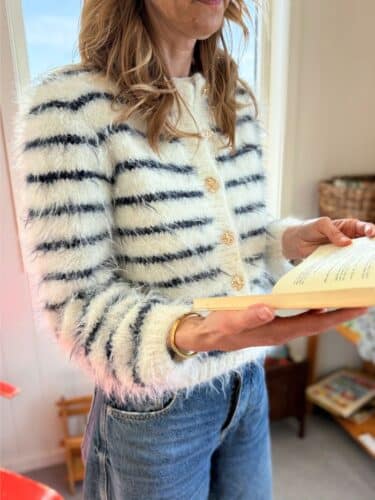 Fuzzy Classic Cardigan fra Tove Lindtein strikket i hvit og marineblå Basta fra Lana Grossa. Står og leser bok.