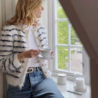 Fuzzy Classic Cardigan fra Tove Lindtein strikket i hvit og marineblå Basta fra Lana Grossa. Sitter i vindusposten og med en kopp.