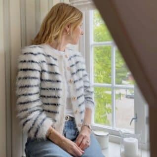Fuzzy Classic Cardigan fra Tove Lindtein strikket i hvit og marineblå Basta fra Lana Grossa. Sitter i vindusposten og ser ut.