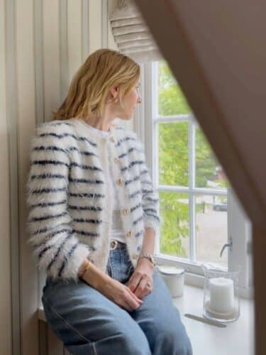 Fuzzy Classic Cardigan fra Tove Lindtein strikket i hvit og marineblå Basta fra Lana Grossa. Sitter i vindusposten og ser ut.